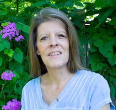 an older woman standing in front of purple flowers and greenery with her eyes closed