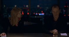 a man and woman sitting at a table in front of a window with the city lights behind them