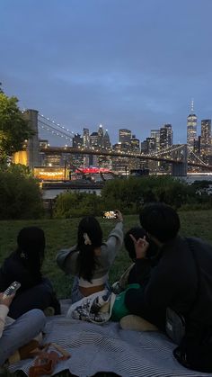 four people sitting on a blanket in front of the city skyline at night, taking pictures with their cell phones