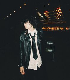 a young man wearing a black leather jacket and white t - shirt is standing in the dark