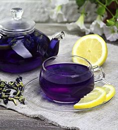 a glass tea pot filled with purple liquid next to lemon slices and a cup of green tea