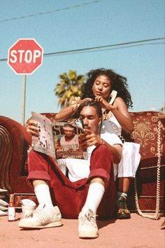 a man and woman sitting on the ground next to a stop sign with a newspaper in front of them