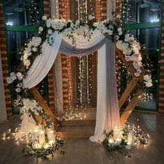 a wedding arch decorated with white flowers and greenery is lit up by fairy lights