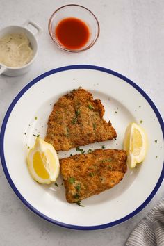 two pieces of fried fish on a white plate with lemon wedges and dipping sauce