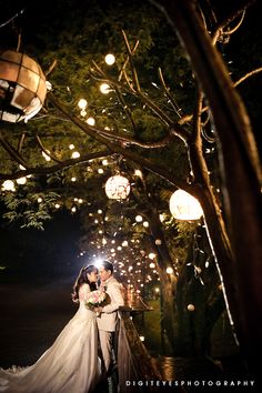 a bride and groom kissing under the lights at night in front of a tree with lanterns