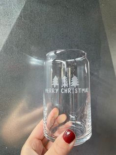 a person holding up a glass with merry christmas written on the side and pine trees behind it