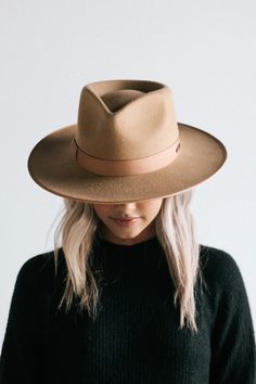 a woman wearing a brown hat and black sweater standing in front of a white wall