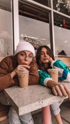 two young women sitting at a table with coffee cups in their hands and one holding her chin up