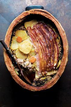 a wooden bowl filled with potatoes and bacon on top of a black table next to a spoon