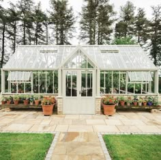 a white greenhouse with potted plants in the front and on the side, surrounded by greenery