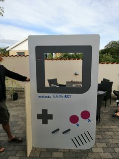 a man standing next to an old fashioned game boy machine on a brick patio with chairs and tables in the background