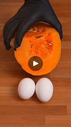 a person in black gloves holding an orange and two eggs on a wooden floor with one hand