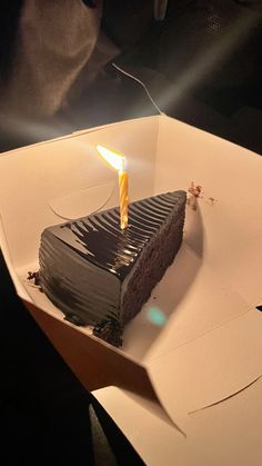 a slice of chocolate cake with a single candle on it sitting in a white box