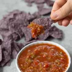 a hand holding a tortilla chip over a bowl of salsa with chips in the background