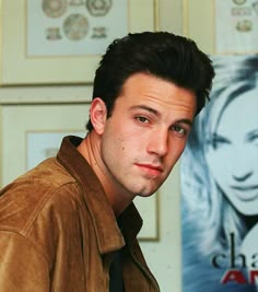 a man in a brown jacket and tie standing next to a wall with posters on it