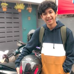 a young man standing next to a motorcycle wearing an orange and blue hoodie with his hands in his pockets