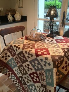 a table with a quilt on it in front of a window and a lamp next to it