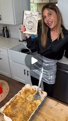 a woman holding up a book in front of a casserole dish on a kitchen counter