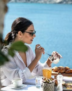 a woman sitting at a table with food and drinks in front of her, overlooking the water