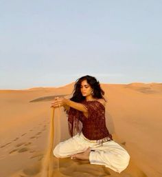 a woman sitting on top of a sand dune holding a stick in her hand and wearing white pants