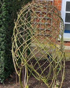 a sculpture made out of branches in front of a brick wall and shrubbery behind it