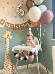 a baby sitting in a high chair next to a giraffe and balloon garland