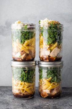 four mason jars filled with pasta, meat and vegetables on a gray tablecloth covered surface