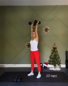 a woman in red pants and white tank top lifting two black dumbbells over her head