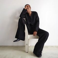 a man sitting on top of a white chair wearing a black suit and tie with his hands behind his head