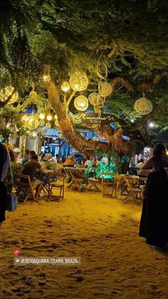 people are sitting at tables under the shade of trees and lanterns hanging from the ceiling