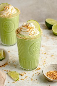 two green drinks with whipped cream and limes on the side next to each other