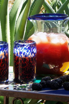 two glasses and pitcher sitting on a table next to some blackberries in front of a potted plant