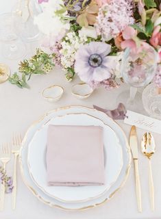 a place setting with pink napkins and goldware on a white table cloth surrounded by flowers