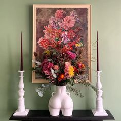 a vase with flowers on a table next to two candles and a painting in the background