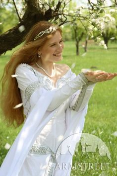 a woman dressed in white is holding out her hand to the side while standing under a tree