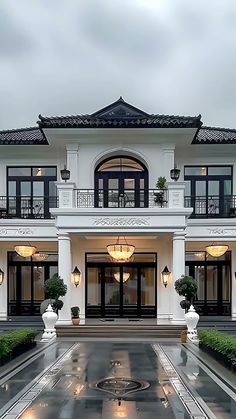 a large white house with lots of windows and lights on the front door, surrounded by greenery