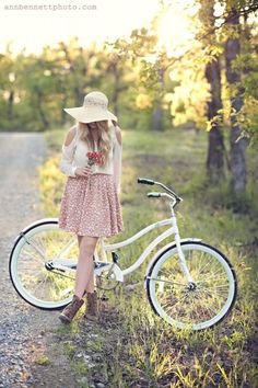 a woman standing next to a white bike on a dirt road in the woods wearing a straw hat