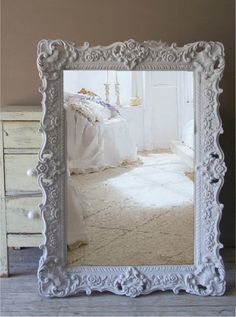 an ornate white frame sits on the floor in front of a dresser and mirror with a bed behind it