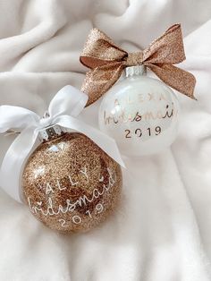 two personalized christmas ornaments on a white blanket with a brown bow and name written on the ornament