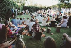 a group of people sitting on top of a grass covered field next to trees and bushes