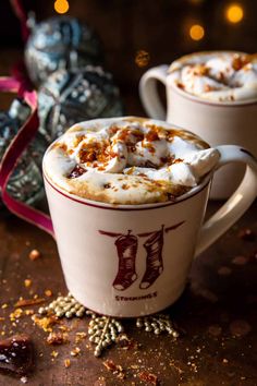 two mugs with hot chocolate and whipped cream in them, on a table next to christmas decorations