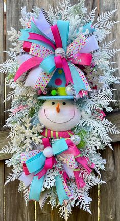 a snowman wreath hanging on the side of a wooden fence with pink, blue and green bows