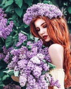 a woman with long red hair holding purple lilacs in her hands and looking at the camera