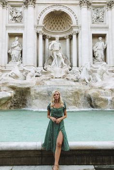 a woman standing in front of a fountain wearing a green dress and tan sandals with her legs crossed