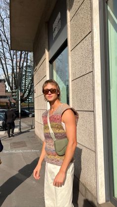 a woman is standing on the sidewalk in front of a building wearing sunglasses and a knitted vest