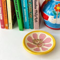 a yellow and pink ring sits in front of books on a white table next to a vase