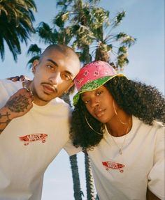 two people standing next to each other with palm trees in the background and one person wearing a pink hat