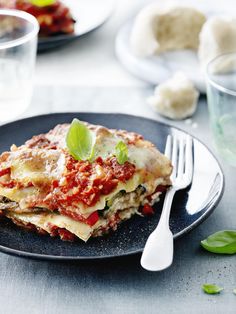 a black plate topped with lasagna covered in sauce and basil next to a glass of water