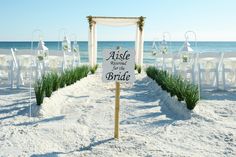 a sign that is on the sand in front of some grass and chairs with an aisle going to the beach