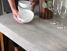 a person holding a bowl on top of a wooden table next to wine glasses and flowers